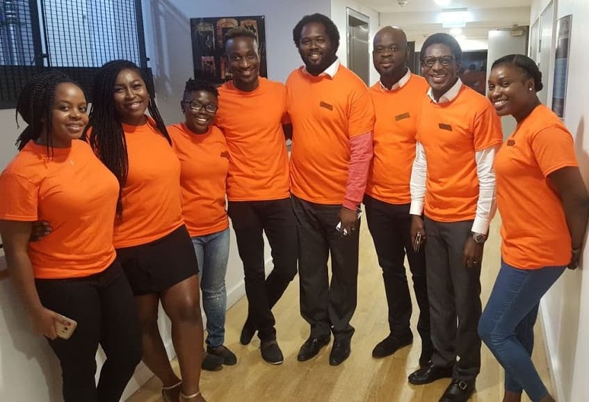 Eight Cee4Bee volunteers in orange team shirts, standing together in a corridor before an event